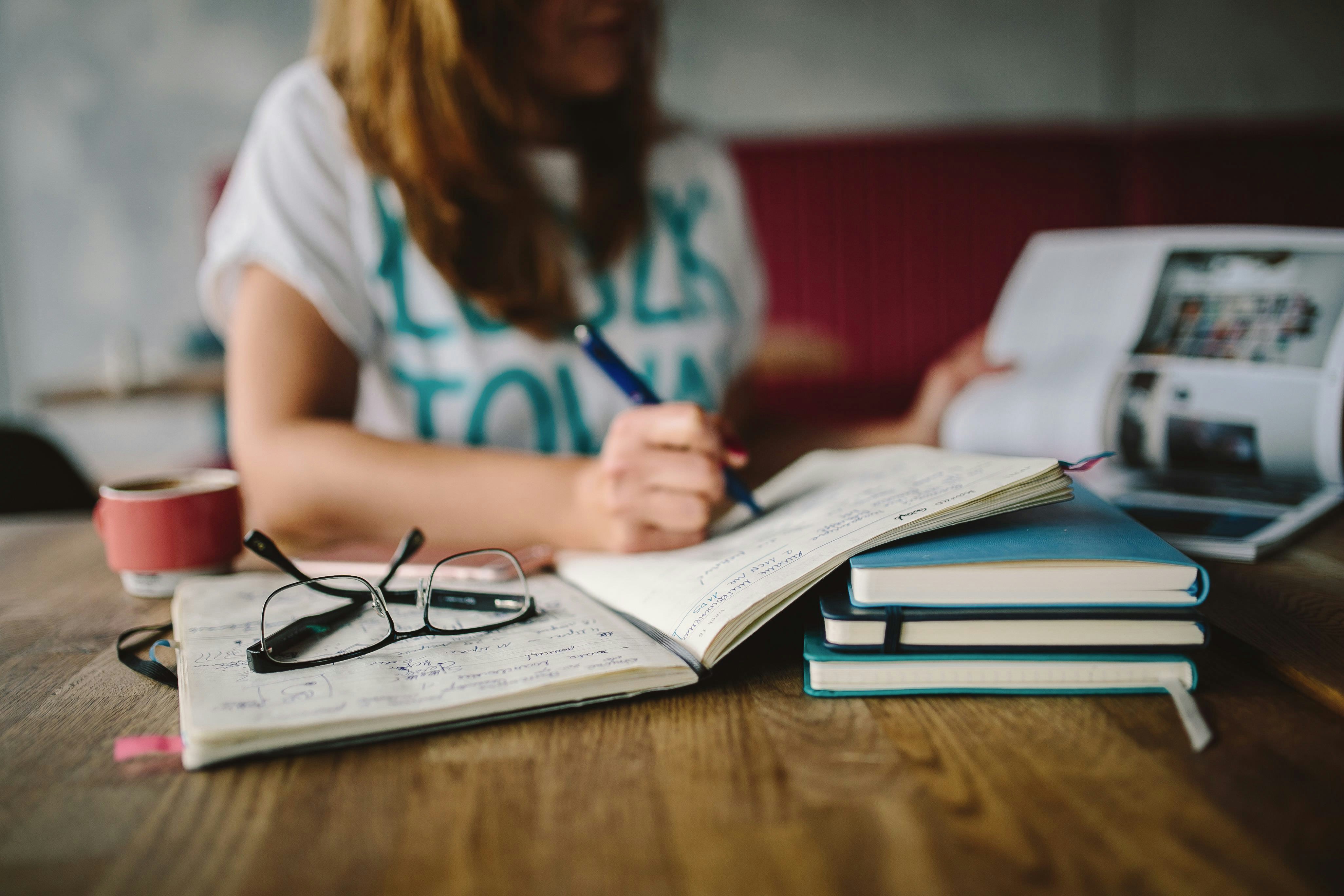 writing in a journal beside glasses and a stack of books