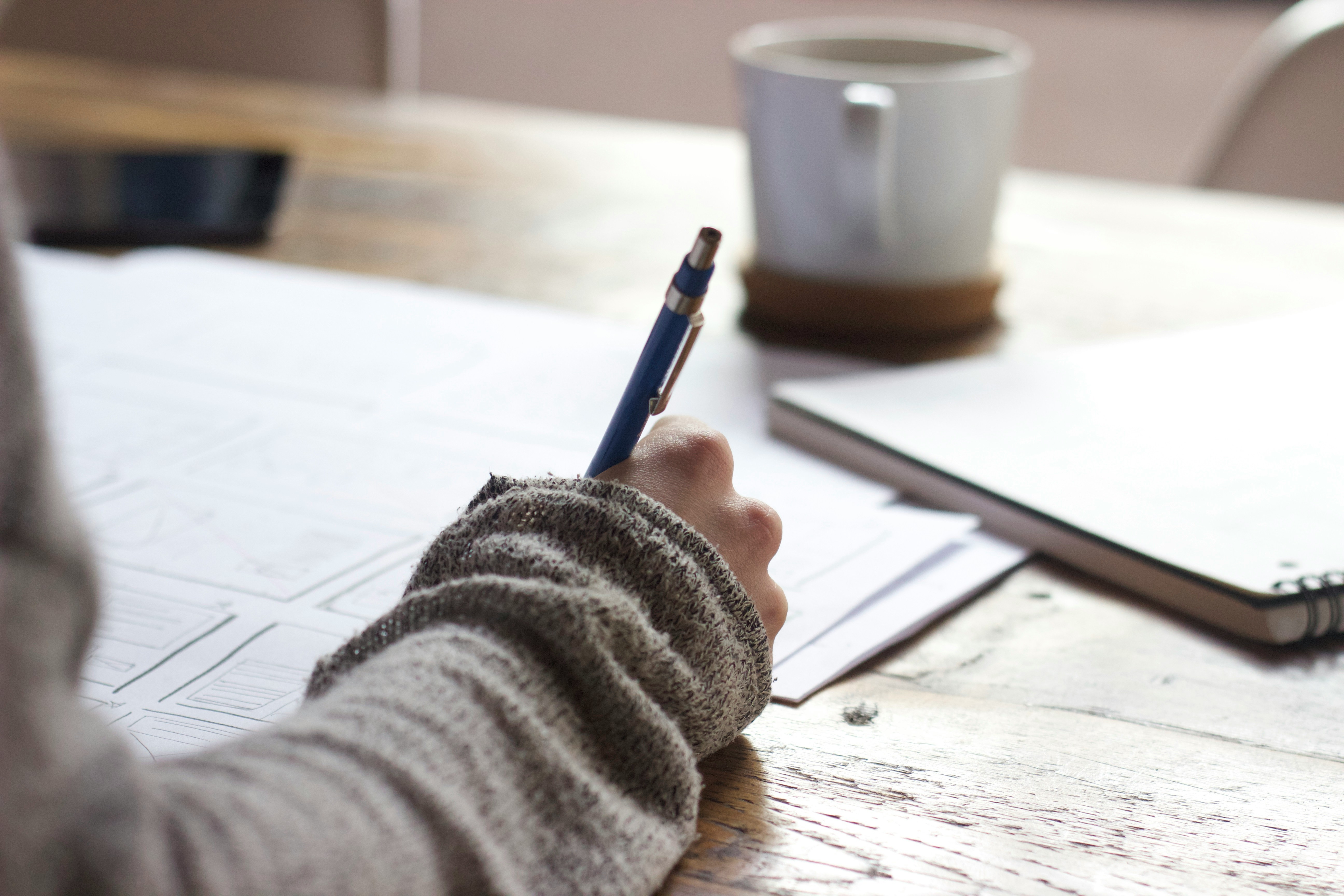 a hand writing at a table beside a mug