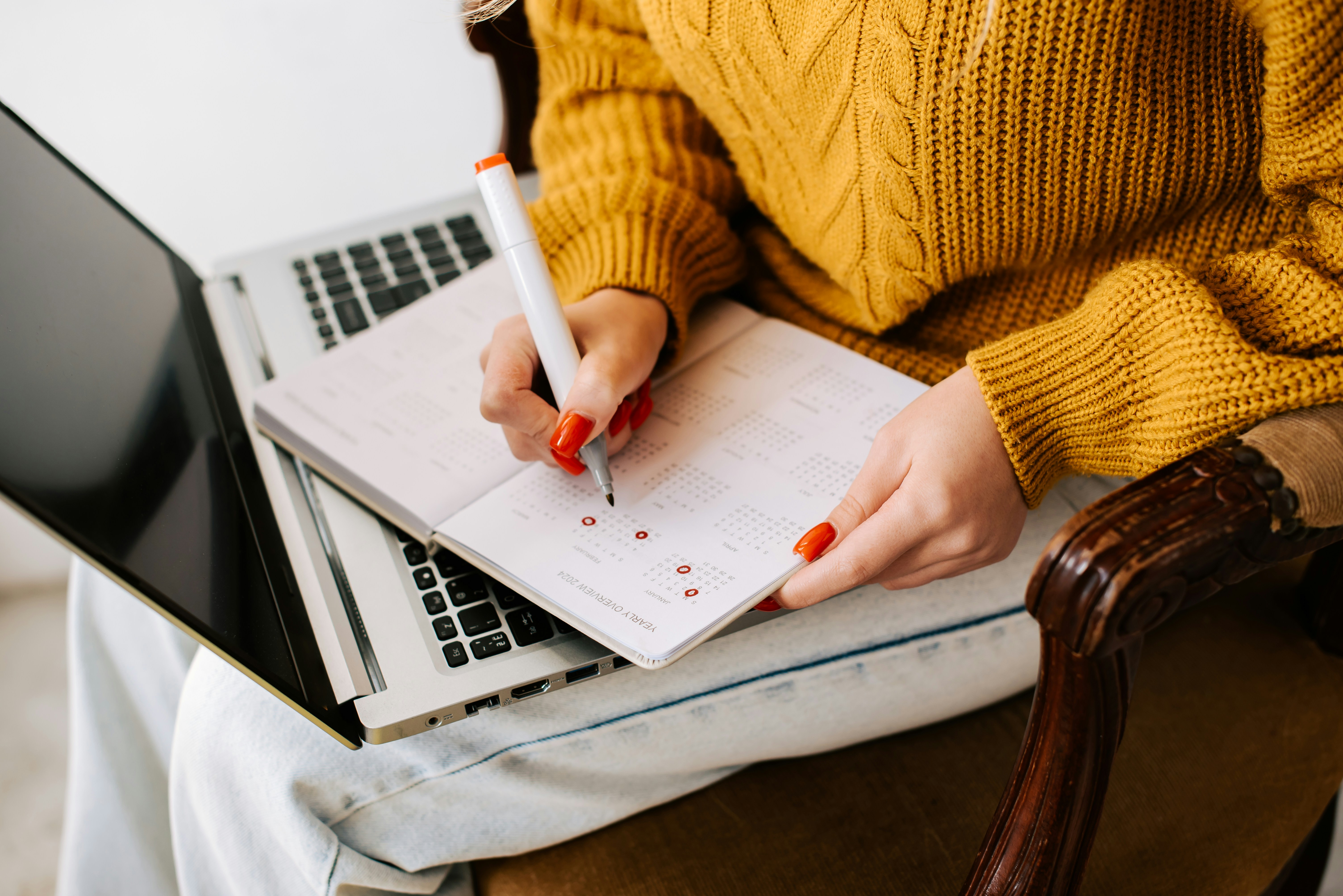 a woman in a yellow jumper writing in a notebook beside a laptop