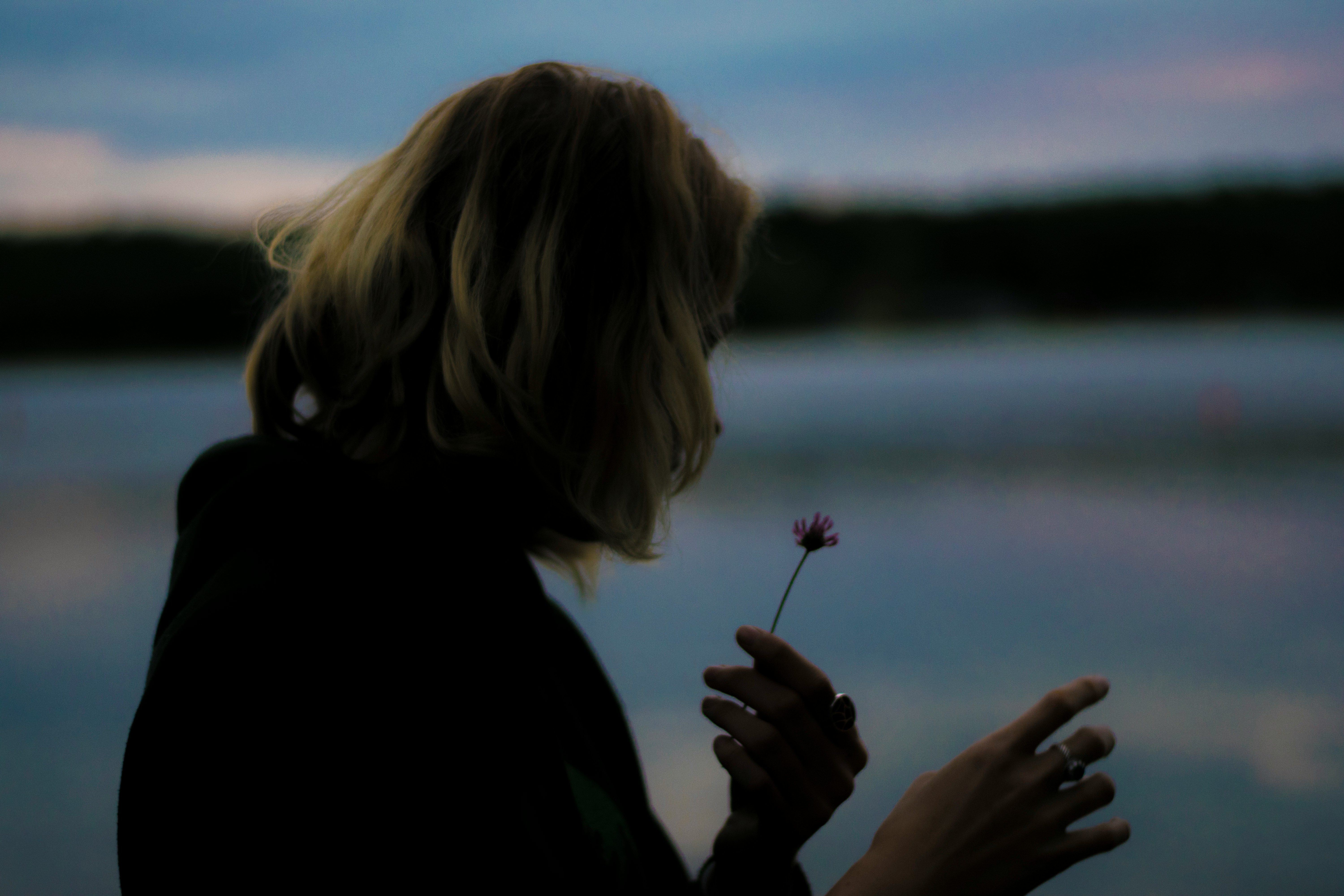 a woman holding a single flower at the water's edge at dusk