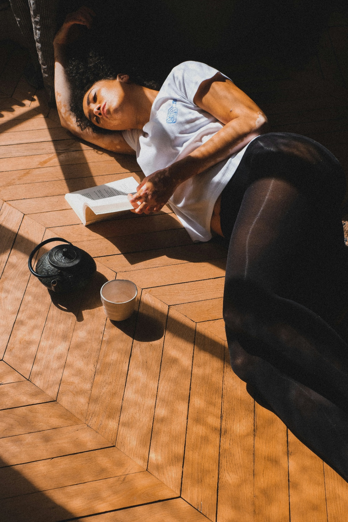 someone lying on a wooden floor reading beside a teapot